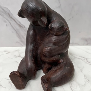 May include: A dark brown bear figurine, carved from wood, depicts a mother bear cradling a cub. The sculpture has a smooth, polished finish, with visible wood grain. The bear is seated, with its head bowed, creating a sense of tenderness.