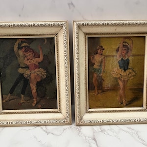 May include: Two framed vintage ballet art prints. Each print features a ballerina in a tutu. The frames are off-white with a decorative border. The ballerina's costumes are pink, blue, and yellow.