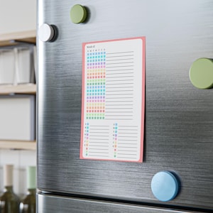 May include: A rectangular, magnetic weekly planner with a pink border is attached to a stainless steel refrigerator. The planner features a grid of colorful squares and lined sections for notes. The text "Week of:" is visible at the top.