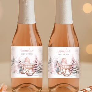 Peut inclure: Deux bouteilles de vin rosé avec des bouchons en feuille blanche et des étiquettes indiquant "Amanda's Baby Shower" avec une illustration de landau. Deux pailles rayées et des fleurs roses sont sur la table.