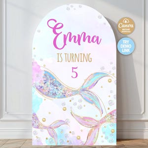 Pode incluir: Uma placa de festa de aniversário com tema de sereia com o nome "Emma" em script rosa, a frase "IS TURNING 5" e ilustrações de caudas de sereia. A placa tem um esquema de cores pastel com bolhas e detalhes dourados.