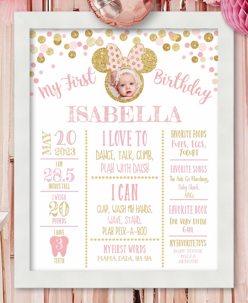 May include: Pink and gold first birthday sign with a photo of a baby girl in a Minnie Mouse headband. The sign includes the baby's name, Isabella, and details about her first year, including her height, weight, and favorite things.