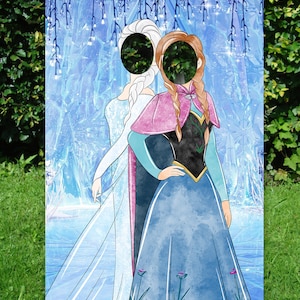 Puede incluir: Un recorte de cart&oacute;n de dos princesas, Elsa y Anna, de la pel&iacute;cula Frozen. El recorte tiene un agujero para una cara en la cabeza de cada princesa. El fondo es una escena de invierno azul y blanco.