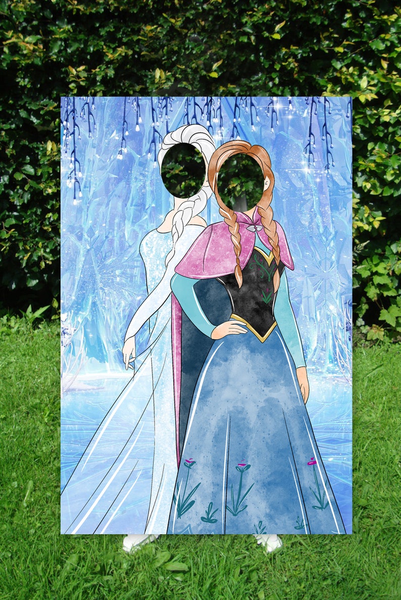 Puede incluir: Un recorte de cart&oacute;n de dos princesas, Elsa y Anna, de la pel&iacute;cula Frozen. El recorte tiene un agujero para una cara en la cabeza de cada princesa. El fondo es una escena de invierno azul y blanco.