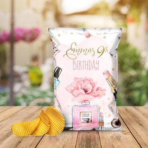 May include: A pink and white chip bag with gold glitter and a watercolor design of a flower, makeup brushes, and perfume bottles. The text "Emma's 9th Birthday" is printed on the bag.