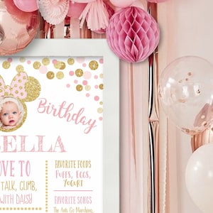 May include: A pink and gold birthday sign with a picture of a baby girl. The sign says "Birthday" and "Ella" in pink and gold letters. It also includes the text "Love to talk, climb, with Daisy", "Favorite Foods: Puffs, Eggs, Yogurt", and "Favorite Songs: The Ants Go Marching".