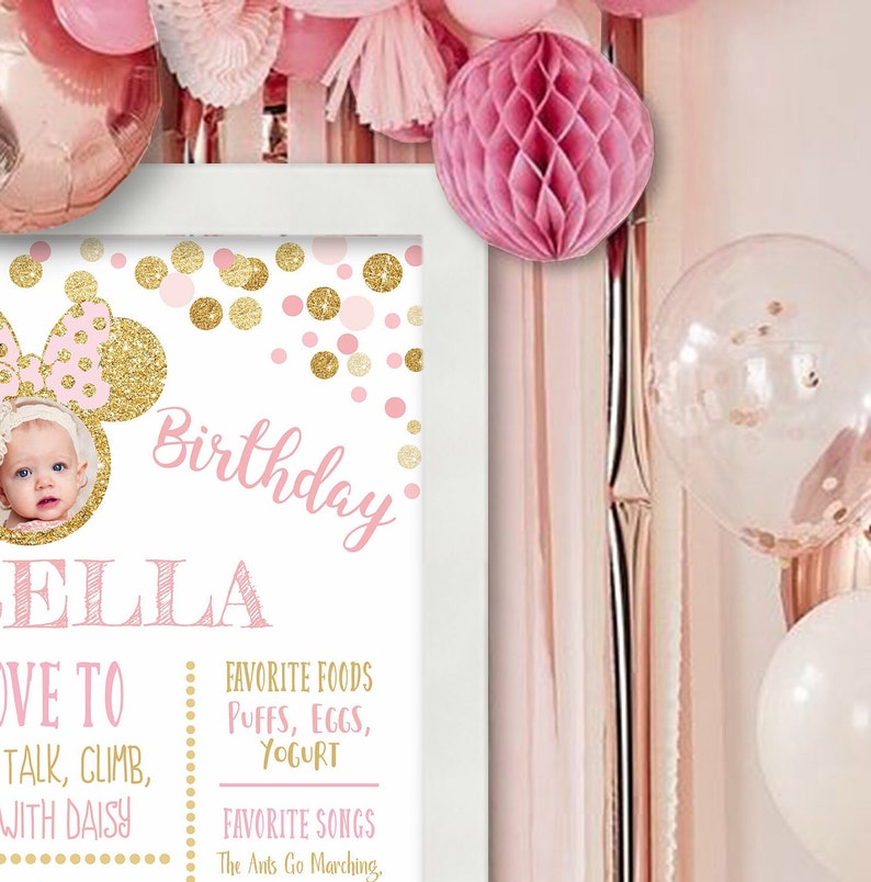 May include: A pink and gold birthday sign with a picture of a baby girl. The sign says "Birthday" and "Ella" in pink and gold letters. It also includes the text "Love to talk, climb, with Daisy", "Favorite Foods: Puffs, Eggs, Yogurt", and "Favorite Songs: The Ants Go Marching".