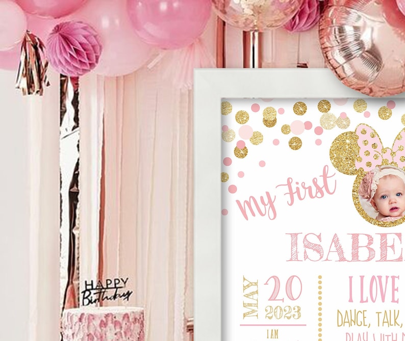 May include: Pink and gold first birthday sign with a photo of a baby girl. The sign says "my first" and "ISABELLA" with a pink and gold Mickey Mouse ears design. The sign also says "MAY 20 2023 I AM" and "I LOVE DANCE, TALK, PLAY WITH MY"