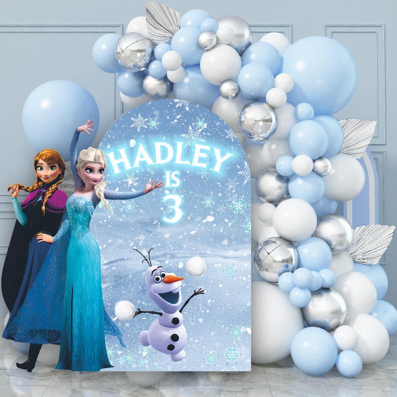 May include: A blue and white backdrop with a Frozen theme featuring Elsa and Olaf. The backdrop has the text "HADLEY IS 3" and is decorated with a blue and silver balloon arch.