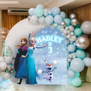 May include: A blue and white Frozen themed party backdrop with a cutout of Elsa and Anna from the movie Frozen. The backdrop also features Olaf, the snowman from the movie. The backdrop is decorated with snowflakes and the text "HADLEY IS 3".