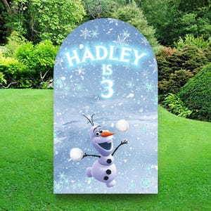 May include: A blue and white Frozen themed birthday party backdrop with a picture of Olaf the snowman throwing snowballs. The backdrop has the text "HADLEY IS 3" on it.