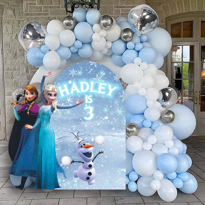May include: A blue and white balloon arch with silver balloons. Two girls dressed as Elsa and Anna from Frozen stand in front of the arch. A sign says "Hadley is 3" with a snowflake design. A cartoon image of Olaf from Frozen is on the sign.