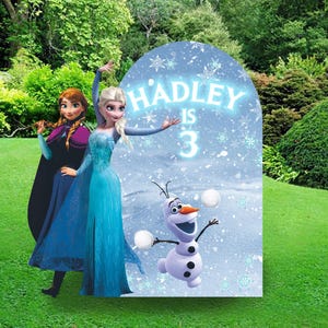 May include: A blue and white Frozen themed birthday party backdrop with a cutout of Elsa and Anna. The backdrop features a snowy scene with Olaf and the text "HADLEY IS 3".