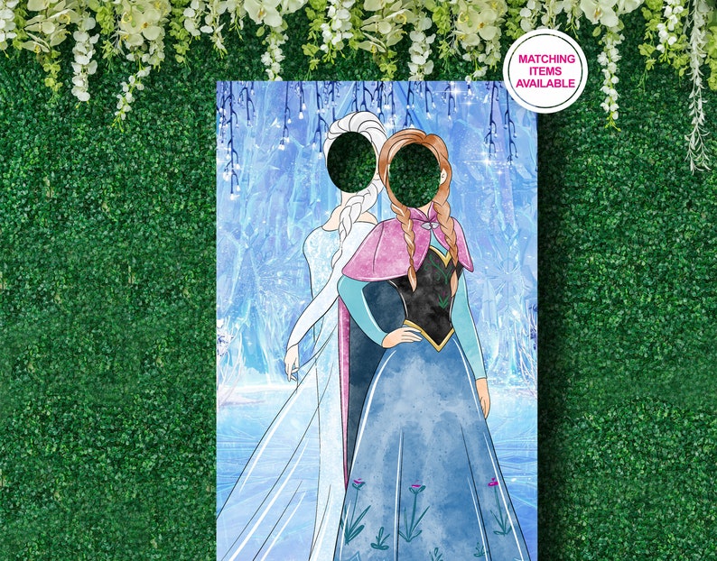 Puede incluir: Un recorte de cart&oacute;n de Elsa y Anna de Frozen, con espacios para las caras. El recorte est&aacute; sobre un fondo azul y blanco con una escena invernal. El texto "Art&iacute;culos a juego disponibles" est&aacute; en un c&iacute;rculo rosa en la esquina superior derecha.