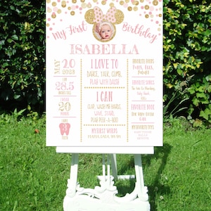 May include: A pink and gold first birthday sign with a photo of a baby girl. The sign includes the baby's name, Isabella, and details about her first year, including her height, weight, and favorite things.