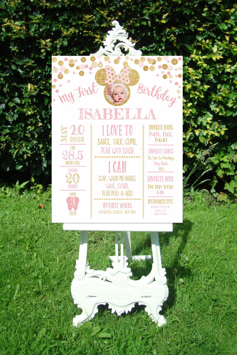 May include: A pink and gold first birthday sign with a photo of a baby girl. The sign includes the baby's name, Isabella, and details about her first year, including her height, weight, and favorite things.