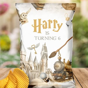 Puede incluir: Una bolsa de patatas fritas blanca con el texto "Harry is Turning 6" en dorado. La bolsa presenta ilustraciones de un castillo, un búho, una llave, un tren y una escoba. Las patatas fritas onduladas están en primer plano.