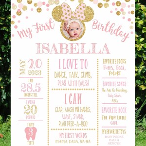 May include: A pink and gold first birthday sign with a photo of a baby girl in a pink and gold Minnie Mouse headband. The sign includes the baby's name, Isabella, and details about her first year, including her height, weight, favorite foods, songs, and toys.