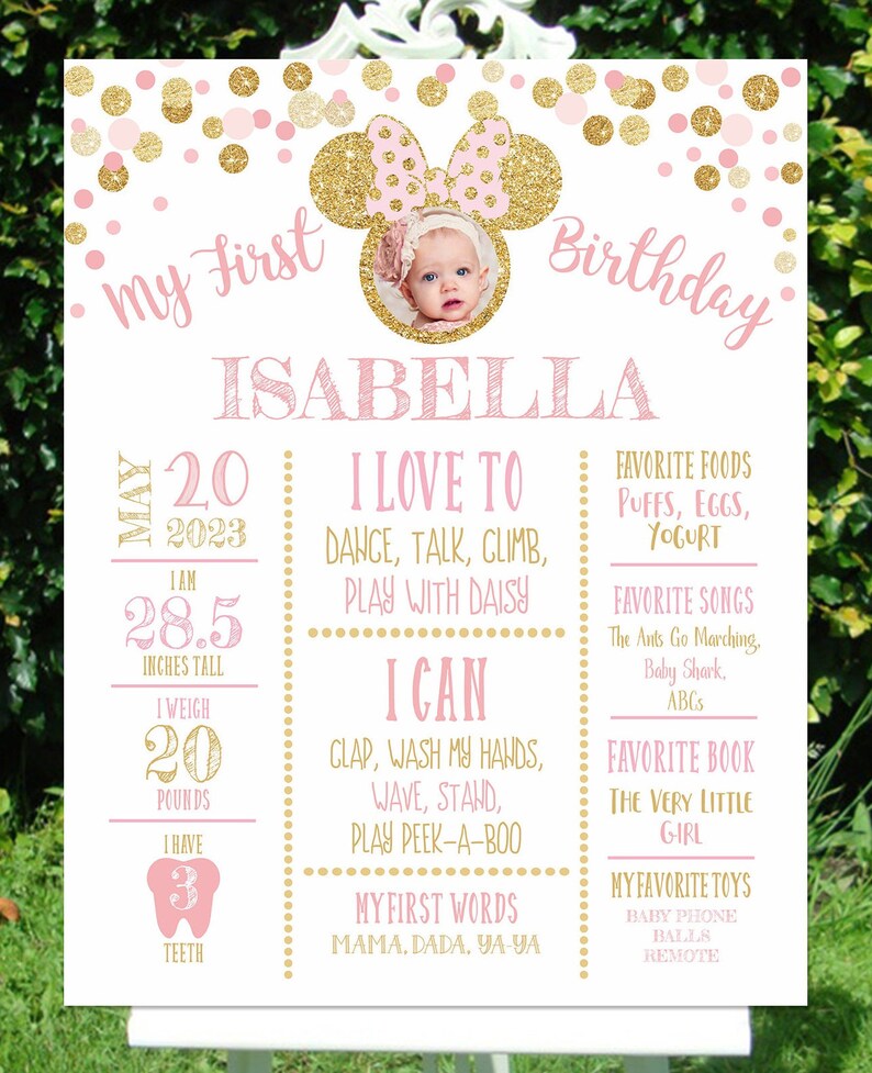 May include: A pink and gold first birthday sign with a photo of a baby girl in a pink and gold Minnie Mouse headband. The sign includes the baby's name, Isabella, and details about her first year, including her height, weight, favorite foods, songs, and toys.