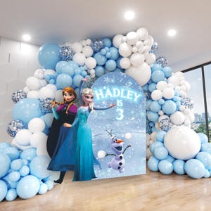May include: A Frozen-themed birthday party backdrop with blue and white balloons, a large cardboard cutout of Elsa and Anna from Frozen, and a sign that says "HADLEY IS 3".