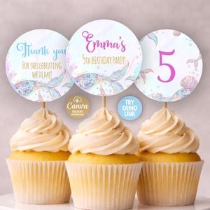 以下が含まれることがあります： 人魚をテーマにしたトッパーが付いた3つのカップケーキ。トッパーは丸く、「Thank you for shellebrating with me!」、「Emma's 5th Birthday Party!」、数字の「5」と書かれています。トッパーには人魚の尾と貝殻のデザインがあります。