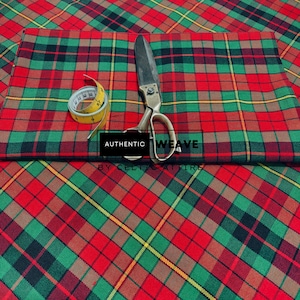 K&ouml;nnte beinhalten: Rot-gr&uuml;ner und schwarzer Stoff mit gelben Akzenten. Ein gelbes Ma&szlig;band und eine Vintage-Schere liegen auf dem Stoff. Der Text "AUTHENTIC WEAVE" und "BY CELTIC ATTIRE" sind sichtbar.