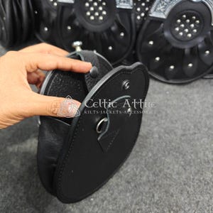Pu&ograve; includere: Sporran in pelle nera con chiusura a patta e borchie argentate. Lo sporran &egrave; aperto, rivelando l'interno. Il testo "Celtic Attire" e "KILTS-JACKETS-ACCESSORIES" &egrave; visibile sullo sporran.