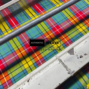 K&ouml;nnte beinhalten: Nahaufnahme eines bunten Tartan-Stoffs in Rot, Gelb, Gr&uuml;n und Blau auf einem Webstuhl. Metall- und wei&szlig;e Holzteile sind zu sehen. Der Text "AUTHENTIC WEAVE BY CELTIC ATTIRE" ist auf dem Stoff abgebildet.