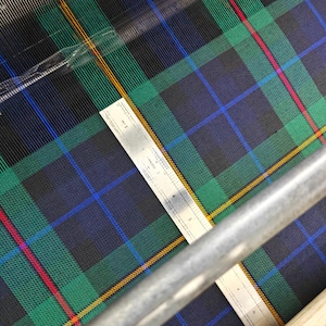 K&ouml;nnte beinhalten: Nahaufnahme eines Tartan-Stoffs mit Lineal. Der Stoff zeigt ein klassisches Karomuster in Gr&uuml;n, Blau, Schwarz, Rot und Gelb. Ein silbernes Lineal, in Zentimetern markiert, liegt auf dem Stoff. Geeignet f&uuml;r Kleidung oder Heimtextilien.