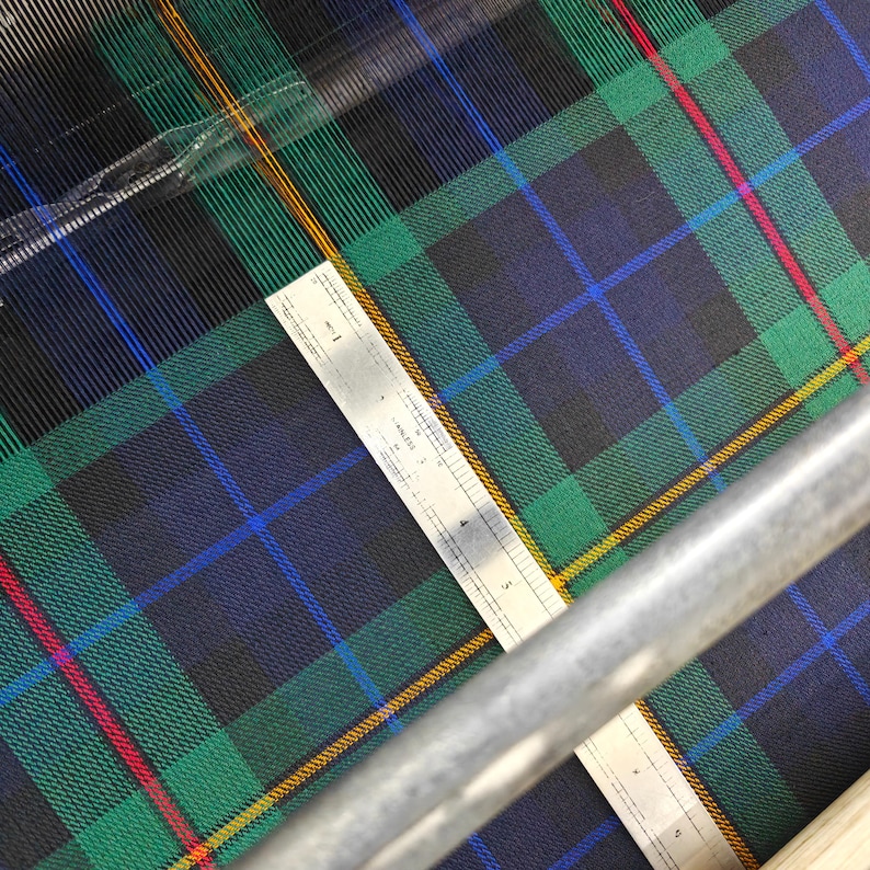 K&ouml;nnte beinhalten: Nahaufnahme eines Tartan-Stoffs mit Lineal. Der Stoff zeigt ein klassisches Karomuster in Gr&uuml;n, Blau, Schwarz, Rot und Gelb. Ein silbernes Lineal, in Zentimetern markiert, liegt auf dem Stoff. Geeignet f&uuml;r Kleidung oder Heimtextilien.