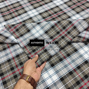 K&ouml;nnte beinhalten: Ein gefalteter Stoff mit Karomuster in Grau-, Schwarz-, Wei&szlig;- und Rott&ouml;nen. Der Stoff hat ein klassisches Karomuster. Der Text "AUTHENTIC WEAVE BY CELTIC ATTIRE" ist auf den Stoff gedruckt.