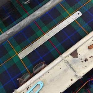 K&ouml;nnte beinhalten: Nahaufnahme eines Tartan-Stoffs in Gr&uuml;n-, Blau- und Rott&ouml;nen, mit einem Metalllineal zur Messung des Stoffs. Eine Brille und ein blaues Werkzeug sind ebenfalls zu sehen.