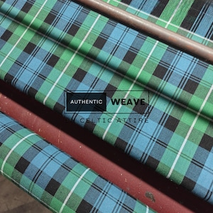 K&ouml;nnte beinhalten: Nahaufnahme eines gefalteten Stoffes mit einem Karomuster in Gr&uuml;n-, Blau-, Schwarz- und Wei&szlig;t&ouml;nen. Der Stoff tr&auml;gt den Text "AUTHENTIC WEAVE BY CELTIC ATTIRE". Der Stoff ist &uuml;ber eine braune Oberfl&auml;che drapiert.