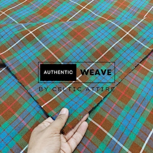 Puede incluir: Primer plano de una tela de cuadros marrón, verde, azul y blanco. La tela está doblada por la mitad y el pliegue es visible en el centro de la imagen. La tela está etiquetada con "AUTHENTIC WEAVE BY CELTIC ATTIRE".