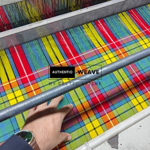 K&ouml;nnte beinhalten: Ein mehrfarbiger Stoff im Webprozess auf einem Webstuhl. Der Stoff weist eine lebendige Mischung aus Rot, Gelb, Blau und Gr&uuml;n auf. Der Text "AUTHENTIC WEAVE BY CELTIC ATTIRE" ist auf dem Stoff zu sehen.