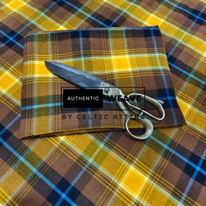 K&ouml;nnte beinhalten: Ein gefalteter Stoff mit Karomuster in Braun-, Gelb- und Blaut&ouml;nen, mit einer offenen Schere darauf. Der Stoff hat ein klassisches Tartanmuster. Der Text "AUTHENTIC WEAVE BY CELTIC ATTIRE" ist sichtbar.