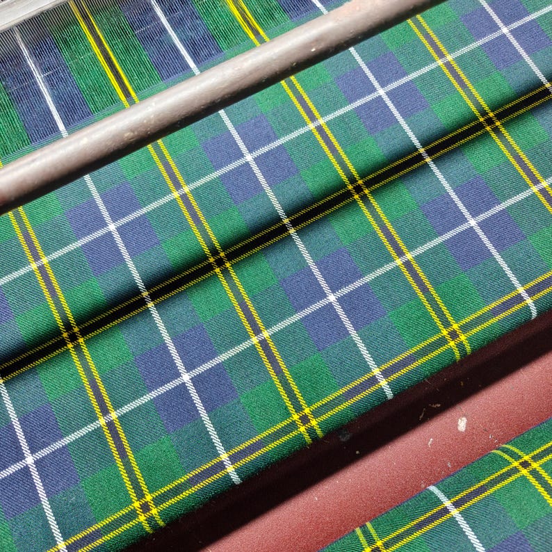 K&ouml;nnte beinhalten: Nahaufnahme eines gr&uuml;n, blau und gelb karierten Stoffes. Der Stoff hat ein sich wiederholendes Muster aus sich kreuzenden Linien, wodurch ein gitterartiges Design entsteht. Der Stoff ist &uuml;ber einer Oberfl&auml;che drapiert, mit einer Metallstange oben sichtbar.