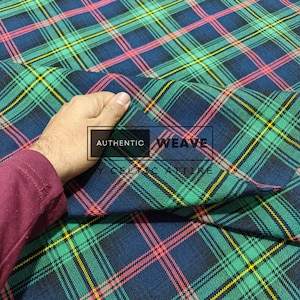 K&ouml;nnte beinhalten: Nahaufnahme eines karierten Stoffes mit einem sich wiederholenden Muster aus sich kreuzenden Linien in Gr&uuml;n, Blau, Gelb und Rosa. Der Stoff wird hochgehalten und zeigt die Textur und das Gewebe. Der Text "AUTHENTIC WEAVE" ist sichtbar.