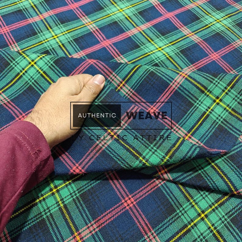 K&ouml;nnte beinhalten: Nahaufnahme eines karierten Stoffes mit einem sich wiederholenden Muster aus sich kreuzenden Linien in Gr&uuml;n, Blau, Gelb und Rosa. Der Stoff wird hochgehalten und zeigt die Textur und das Gewebe. Der Text "AUTHENTIC WEAVE" ist sichtbar.