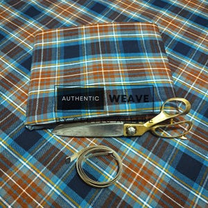 K&ouml;nnte beinhalten: Gefalteter Stoff mit blauem, braunem und wei&szlig;em Karomuster und dem Text "AUTHENTIC WEAVE". Eine goldfarbene Schere und ein Ma&szlig;band liegen auf dem Stoff.