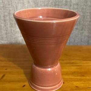 May include: A vintage, dusty rose-colored ceramic vase with a flared top and a pedestal base. The vase has horizontal ridged detailing around the middle. The piece is likely from the mid-20th century and is a decorative item.
