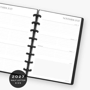 Op de afbeelding: Een planner met schijfbinding open op een weekoverzicht voor november 2027. De witte pagina's bevatten zwarte tekst, horizontale lijnen voor dagelijkse planning en een notitiesectie. Een zwarte plastic schijfrug houdt de pagina's bij elkaar. Een ronde zwarte badge linksonder vermeldt 2027 HALF LETTER SIZE.