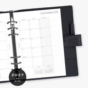 May include: A black A5 size planner with a six-ring metal binder mechanism. The open page displays a monthly calendar for November 2027 with grid lines, a pen loop on the right side, and a circular black graphic overlay stating 2027 A5 SIZE.