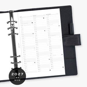 Peut inclure: Un classeur agenda noir format A5 pour 2027 avec une couverture texturée et un mécanisme métallique argenté à six anneaux. L'agenda ouvert présente une feuille blanche avec une mise en page verticale pour juillet, août et septembre 2027.