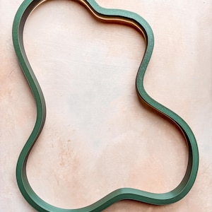 Op de afbeelding: Een onregelmatig gevormd frame met een matte donkergroene buitenkant en een gladde goudkleurige binnenrand. Het frame heeft een doorlopende, vloeiende omtrek met meerdere bochten en inkepingen, waardoor een hol midden ontstaat.