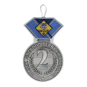 May include: A silver-colored embroidered patch in the shape of a medal. The medal reads "PACK PINEWOOD DERBY CHAMPION" and "2ND" with a laurel wreath. A blue and yellow ribbon with a wolf logo is attached.