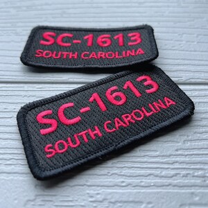 Puede incluir: Dos parches rectangulares negros bordados con el texto "SC-1613 SOUTH CAROLINA" en rosa brillante. Los parches tienen un borde negro y se muestran sobre una superficie gris claro.