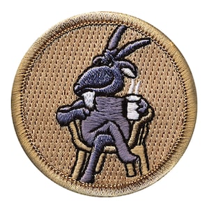 May include: A brown embroidered patch with a purple goat sitting in a chair and holding a cup of coffee. The goat has a white beard and a big smile.