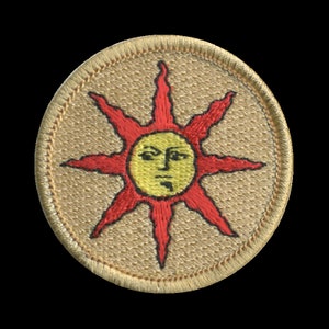 May include: A tan embroidered patch with a red sun with a yellow face and 8 points. The sun has a frowning expression.