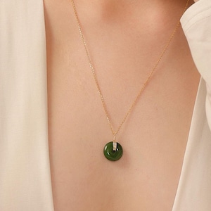 May include: A gold chain necklace featuring a circular dark green jade pendant. The pendant is suspended from a small gold bail attached to the delicate gold link chain. The necklace rests against a plain white long-sleeved garment.
